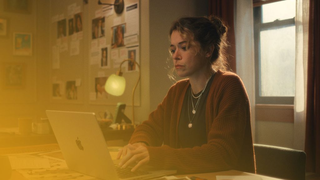 Eine Frau mit hochgesteckten Haaren sitzt konzentriert an einem Schreibtisch und arbeitet an einem Laptop, beleuchtet von einer grünen Lampe und warmem Fensterlicht.
