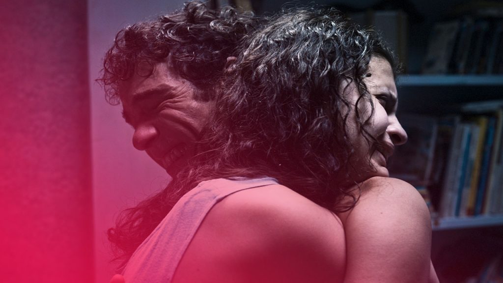 Zwei Personen mit dunklen, lockigen Haaren umarmen sich innig in einem schwach beleuchteten Raum. Beide haben die Augen geschlossen und zeigen einen schmerzverzerrten Gesichtsausdruck. Im Hintergrund ist ein Bücherregal erkennbar.