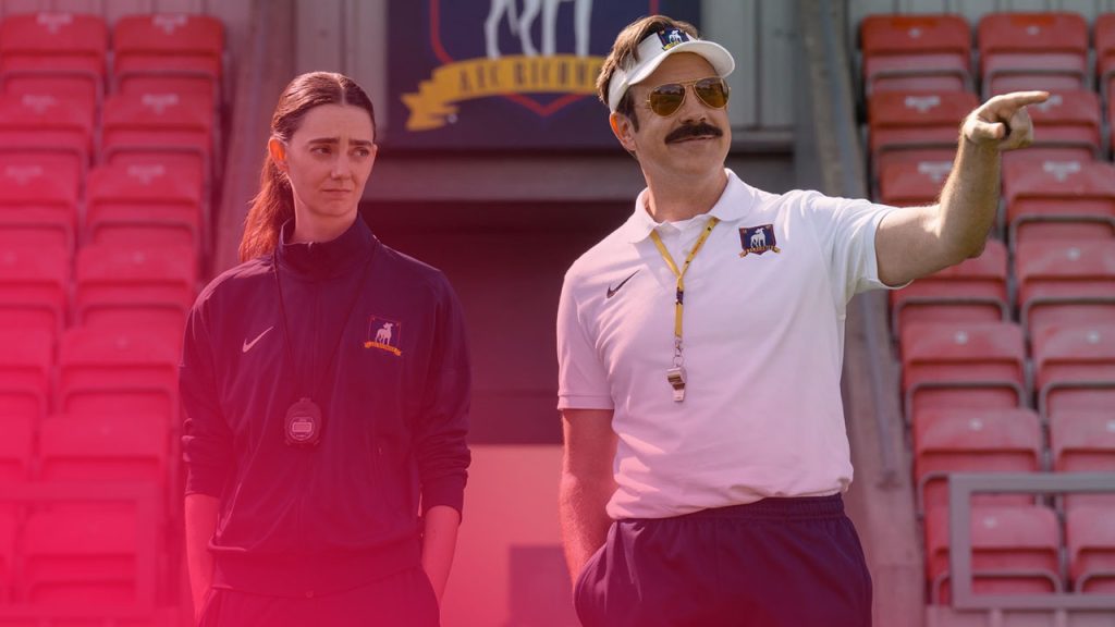 Zwei Trainer in Sportkleidung stehen vor roten Tribünensitzen. Der Mann mit Schnurrbart und Visiermütze zeigt nach rechts, während die Frau neben ihm eine Stoppuhr um den Hals trägt und ernst blickt.