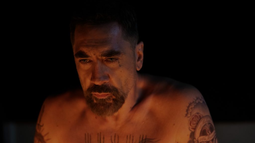 Ein muskulöser, oberkörperfreier Mann mit Bart und Tattoos blickt intensiv in die Kamera, beleuchtet von warmem Licht vor schwarzem Hintergrund.