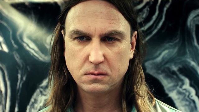 Mann mit langen dunklen Haaren und intensivem Blick, blaue Augen, ernster Gesichtsausdruck vor einem abstrakten schwarz-weißen Hintergrund mit welligen Mustern.