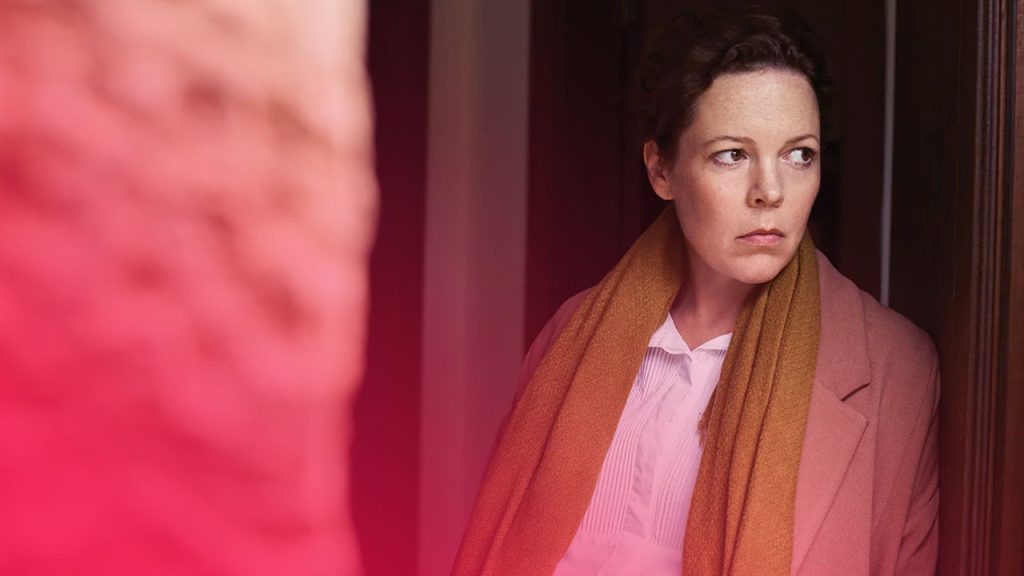 Eine Frau mit hochgesteckten Haaren, bekleidet mit einem rosafarbenen Mantel und einem senfgelben Schal, steht an einen Türrahmen gelehnt und blickt mit ernstem Ausdruck nach links.