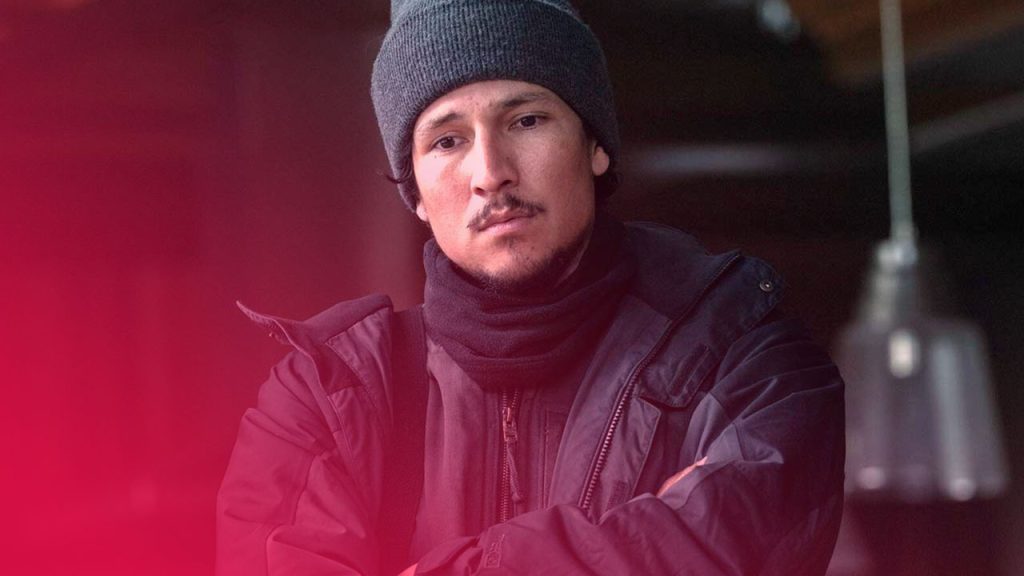 Junger Mann mit dunkler Strickmütze, Schal und Jacke steht mit verschränkten Armen da und blickt ernst in die Kamera; der Hintergrund ist unscharf und zeigt eine hängende Lampe in einem Innenraum.