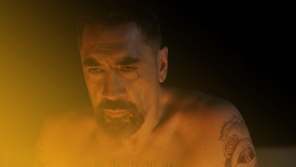 Ein muskulöser, oberkörperfreier Mann mit Bart und Tattoos blickt intensiv in die Kamera, beleuchtet von warmem Licht vor schwarzem Hintergrund.