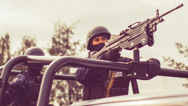 Zwei bewaffnete Polizisten in schwarzer Uniform und Helm stehen auf einem Fahrzeug; einer zielt mit einem Maschinengewehr, der andere im Hintergrund, beide mit vermummten Gesichtern.