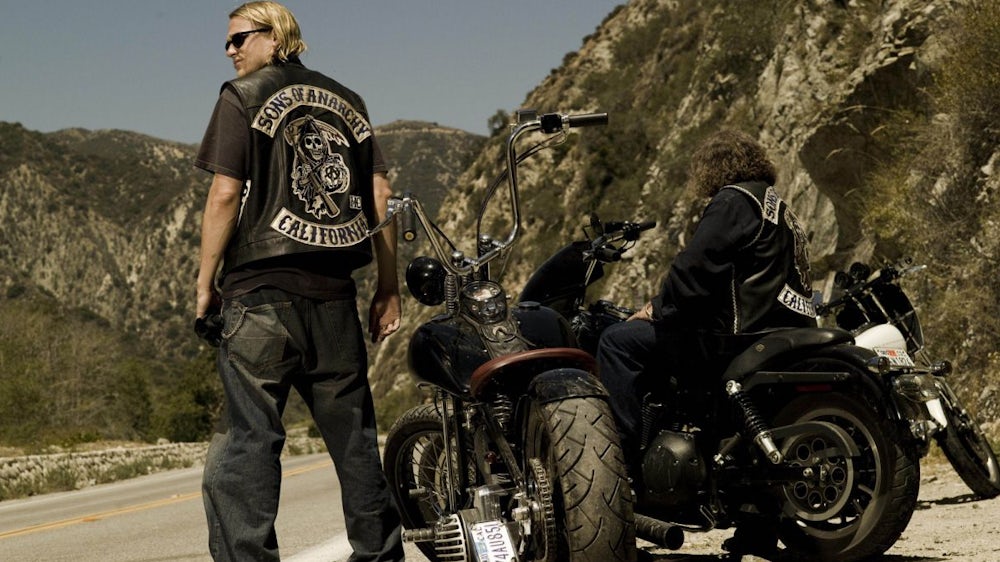 Zwei Männer in Lederwesten mit dem Aufdruck „Sons of Anarchy California“ stehen neben ihren Motorrädern an einer kurvigen Bergstraße, wobei einer der Männer auf sein Motorrad steigt und der andere mit dem Rücken zur Kamera steht.