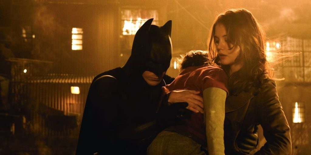 Batman und eine Frau stehen im warmen Schein einer nächtlichen Stadt und halten gemeinsam ein Kind schützend im Arm.