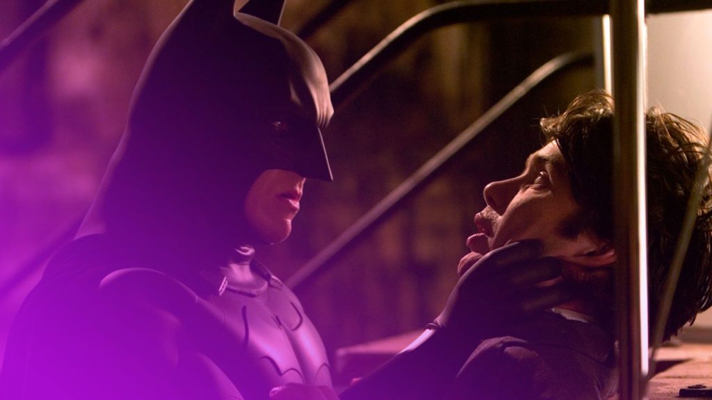 Batman in seinem dunklen Kostüm hält einen Mann am Kragen gepackt und drückt ihn gegen ein Metallgeländer in einer düsteren, schattigen Umgebung.