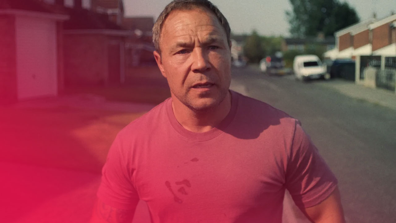 Mann in rosafarbenem, verschmutztem T-Shirt mit Tattoo am Arm steht auf einer Wohnstraße, im Hintergrund Häuser und Autos.