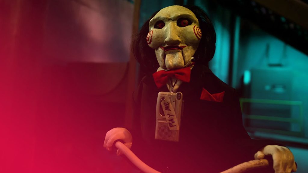 Gruselige Billy-Puppe auf einem Dreirad mit roter Fliege und Diktiergerät „Play Me“ in einem düsteren, grünlichen Raum.