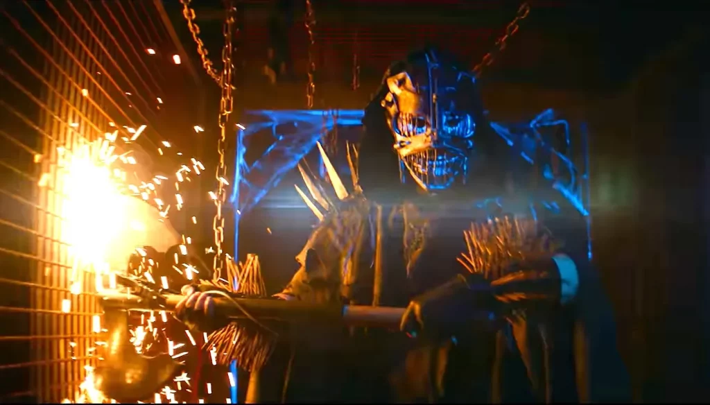 Eine Person in einer metallischen, gesichtsbedeckenden Maske mit spitzen Zähnen und Hörnern hält eine Funken sprühende Waffe. Die Szene ist dunkel, beleuchtet von blauem und orangefarbenem Licht, mit Ketten und Gittern im Hintergrund. Die Atmosphäre ist bedrohlich und actiongeladen.