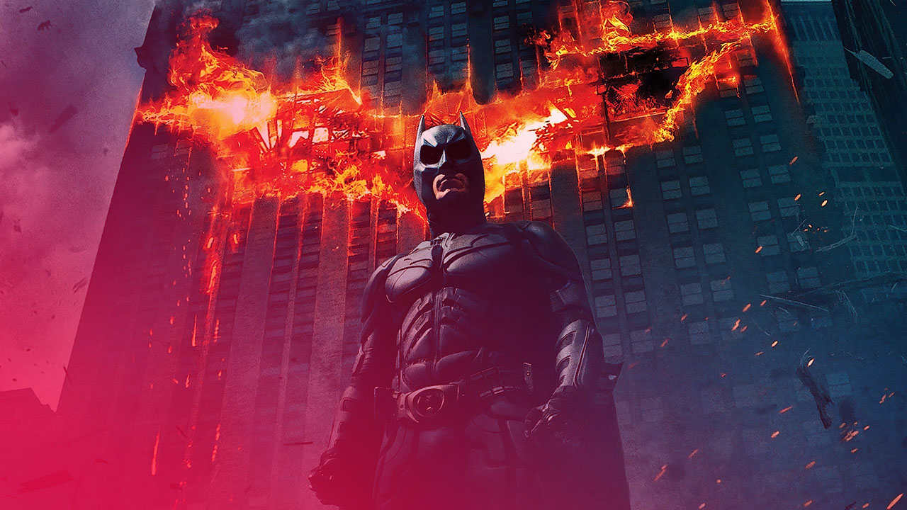 Batman steht vor einem brennenden Hochhaus, dessen Flammen die Form eines Fledermaus-Symbols bilden, während Funken durch die dunkle, rauchige Szene fliegen.