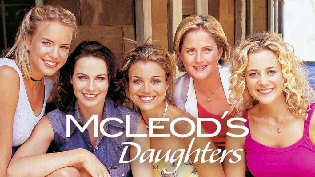 Fünf lächelnde Frauen posieren eng beieinander im Freien vor einem Holzgebäude. Sie tragen legere Sommerkleidung in Weiß, Blau, Rosa und Lila. Der weiße Schriftzug „MCLEOD'S Daughters" ist über dem Bild platziert.