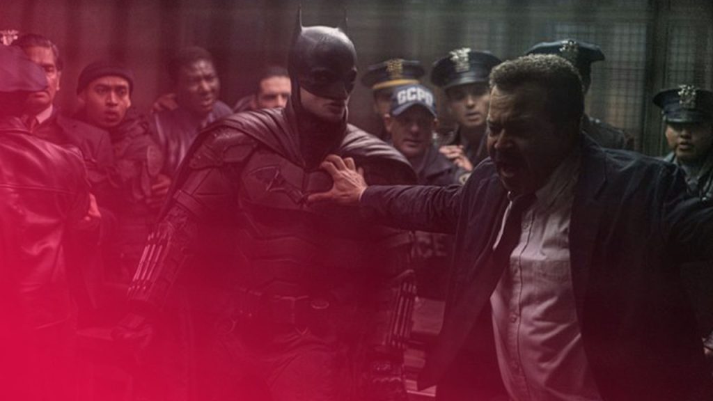 Eine dramatische Szene in einem dunklen, gedämpften Raum, in der Batman in seiner schwarzen Rüstung mit spitzem Helm in der Mitte steht. Ein aufgebrachter, älterer Mann mit Schnurrbart in einem Anzug mit offenem Hemd und einer blauen Krawatte greift ihn am Arm, während er mit offenem Mund schreit. Eine Gruppe von Polizisten in Uniform und Zivilisten steht im Hintergrund und schaut schockiert zu. Der Raum hat eine industrielle Atmosphäre mit sichtbaren Rohren und einem Gitterfenster. Die Beleuchtung ist spärlich und erzeugt eine bedrohliche Stimmung.