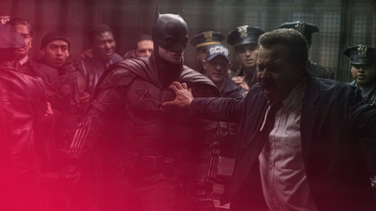 Eine dramatische Szene in einem dunklen, gedämpften Raum, in der Batman in seiner schwarzen Rüstung mit spitzem Helm in der Mitte steht. Ein aufgebrachter, älterer Mann mit Schnurrbart in einem Anzug mit offenem Hemd und einer blauen Krawatte greift ihn am Arm, während er mit offenem Mund schreit. Eine Gruppe von Polizisten in Uniform und Zivilisten steht im Hintergrund und schaut schockiert zu. Der Raum hat eine industrielle Atmosphäre mit sichtbaren Rohren und einem Gitterfenster. Die Beleuchtung ist spärlich und erzeugt eine bedrohliche Stimmung.