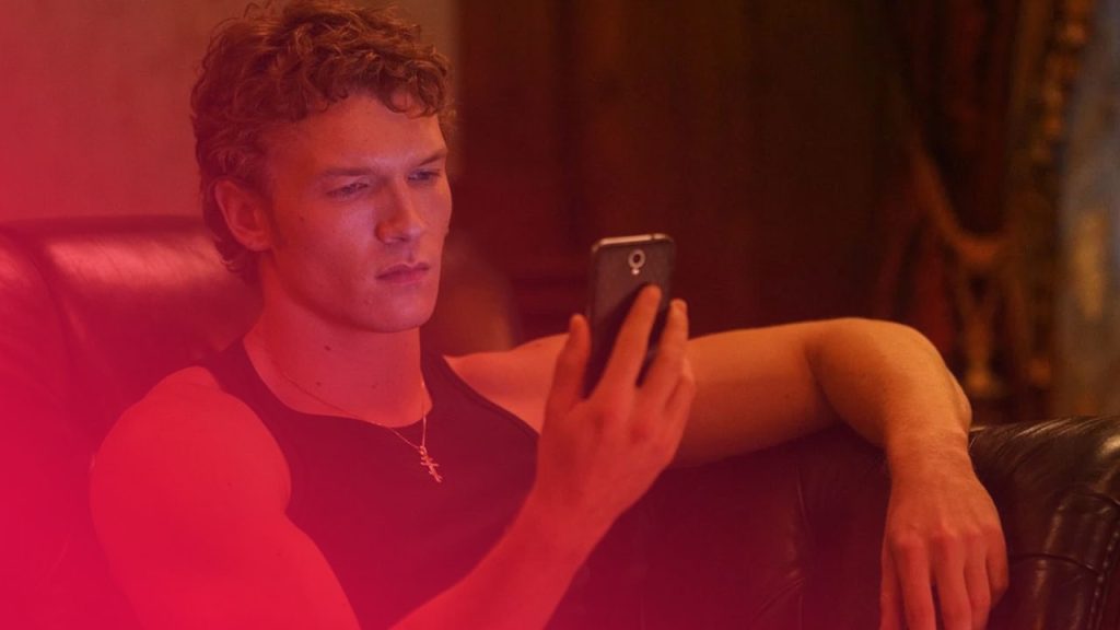 Junger Mann mit lockigen Haaren und schwarzem Tanktop sitzt auf schwarzem Ledersofa und blickt konzentriert auf sein Smartphone.