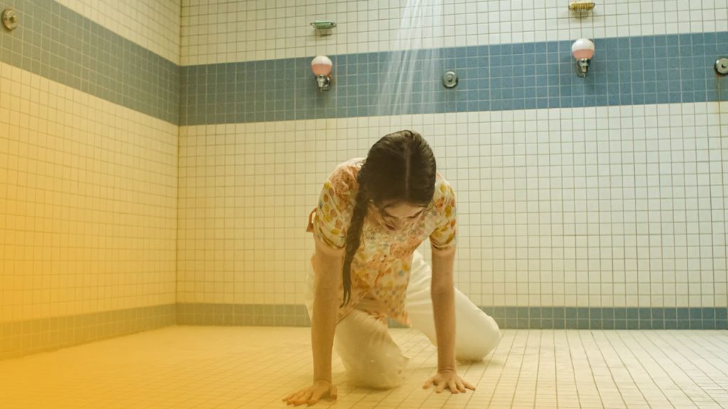 Eine junge Frau mit Zopf kniet auf allen Vieren auf gefliestem Boden eines Duschraums, während Wasser von oben auf sie herabfällt.