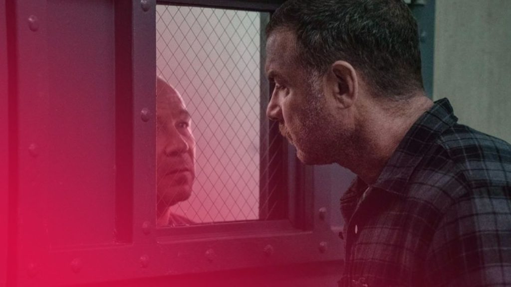 Zwei Männer blicken sich durch das vergitterte Fenster einer schweren Stahltür intensiv an; der Mann im Vordergrund trägt ein kariertes Hemd, der Mann im Hintergrund ist nur teilweise sichtbar.