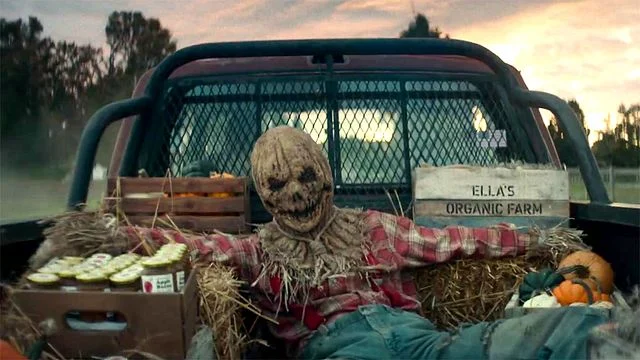 Vogelscheuche im karierten Hemd sitzt auf Pickup-Ladefläche mit Heu, Kürbissen und Kisten, Schild „Ella's Organic Farm", Bäume bei Dämmerung.