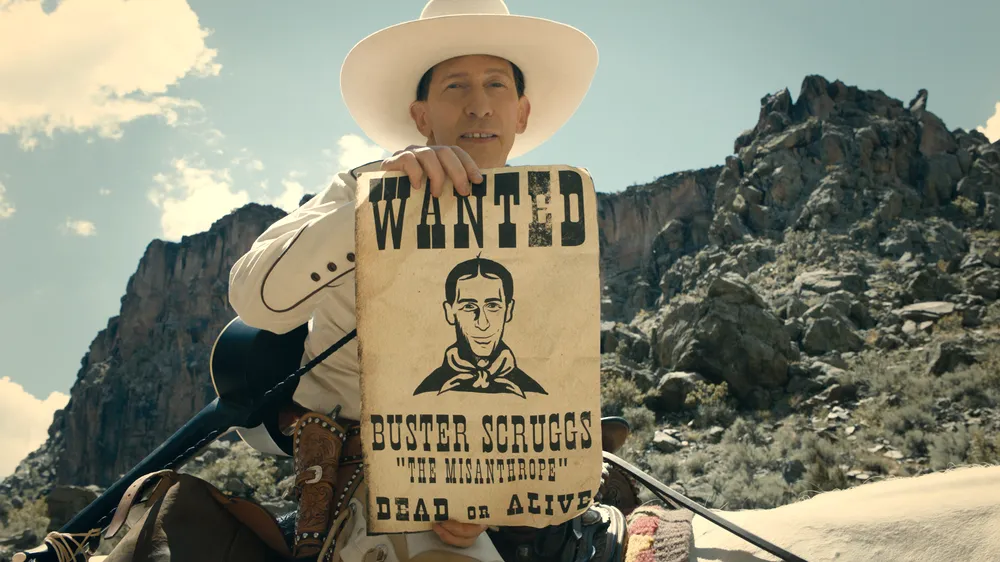 Cowboy mit weißem Hut hält ein Wanted-Plakat mit Porträt und dem Namen Buster Scruggs vor felsiger Wüstenlandschaft.